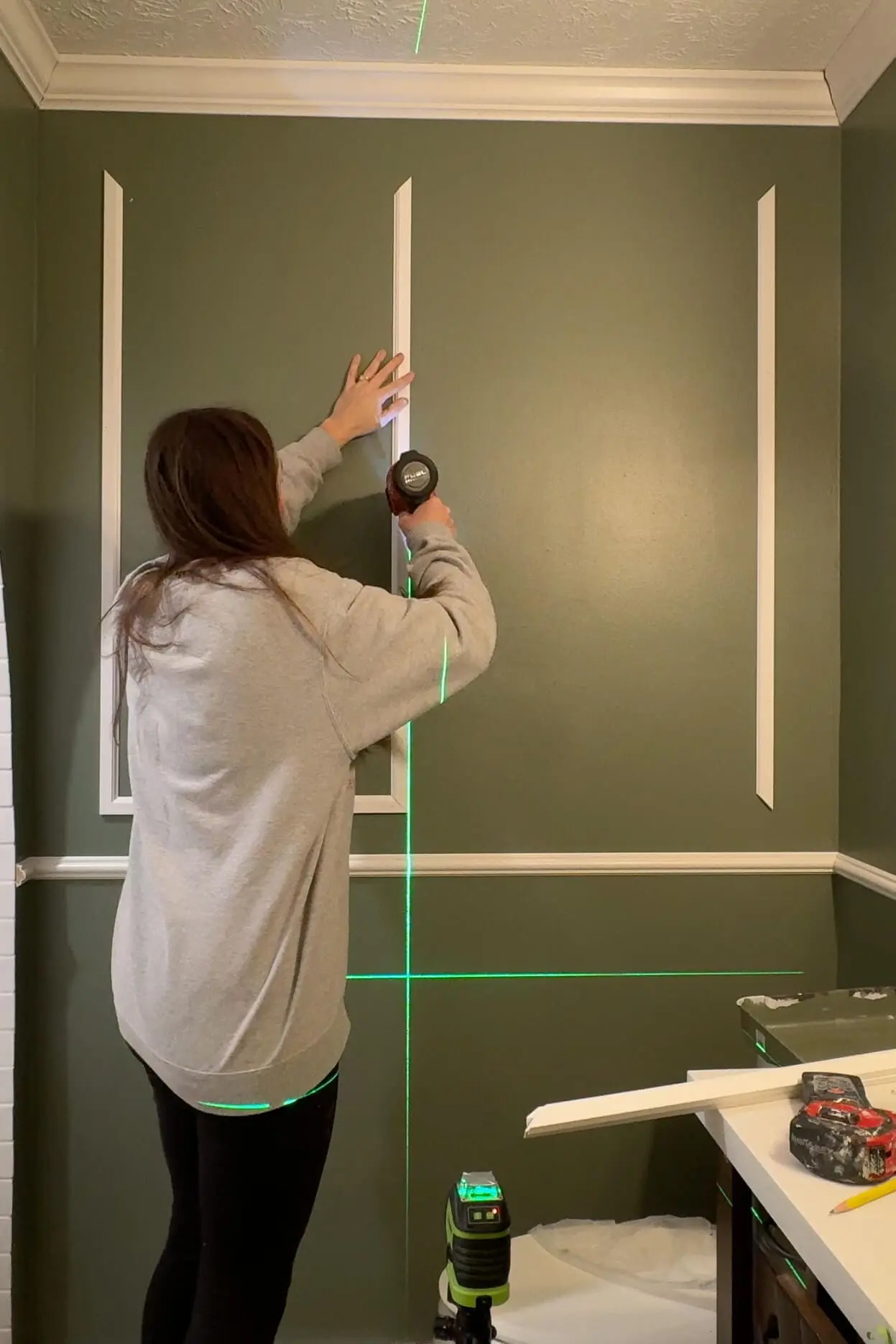 DIY enthusiast using a nail gun to secure trim pieces on a dark green bathroom wall with the aid of a laser level, crafting a sophisticated trim accent wall design.